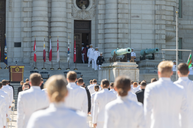 Gruppe von Menschen in weißen Marineuniformen, die auf einer Treppe vor einem neoklassizistischen Gebäude mit Säulen, einem Podium und Flaggen während einer Abschlussfeier stehen.