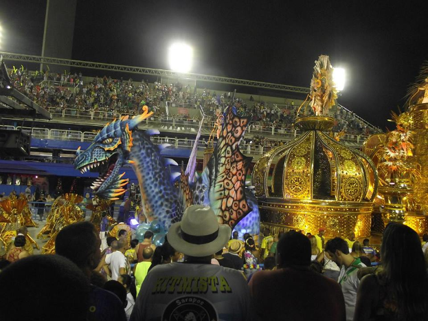 Eine große Menschenmenge umgibt ein hell erleuchtetes Karnevalswagen, der mit farbenfrohen Kostümen geschmückt ist, mit einem Stadion voller Menschen und dem Himmel im Hintergrund.