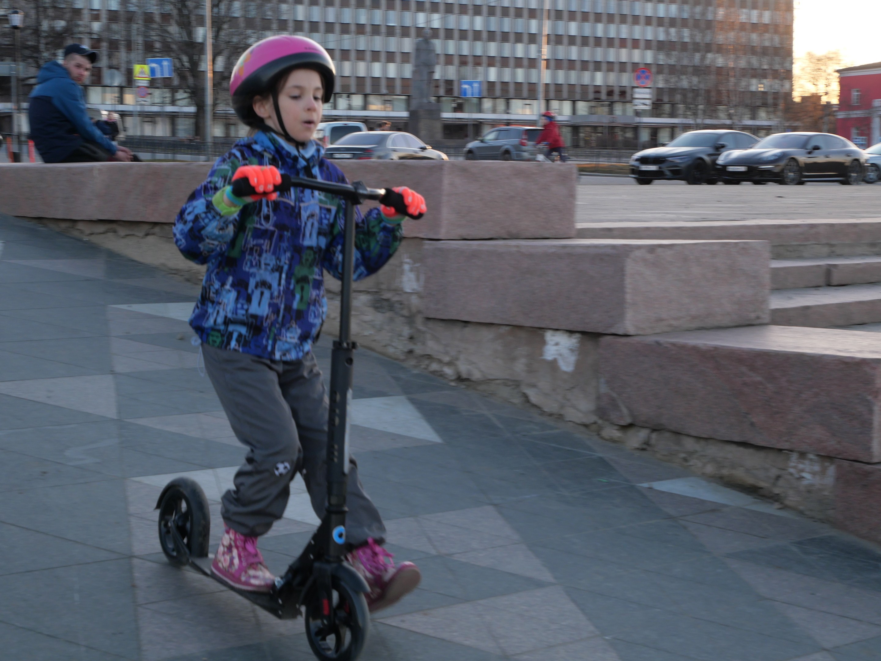Ein junger Junge in einem Helm und Handschuhen fährt einen Roller eine Treppe hinunter, mit Autos, Menschen, Bäumen, Pfosten, Brettern, Gebäuden und einem klaren blauen Himmel im Hintergrund.