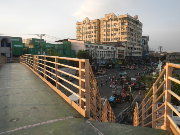 Fußgängerbrücke mit Geländern, die eine vielbefahrene Straße mit Fahrzeugen und Fußgängern überspannt, mit Gebäuden, Bäumen, Strommasten und einem bewölkten Himmel im Hintergrund.