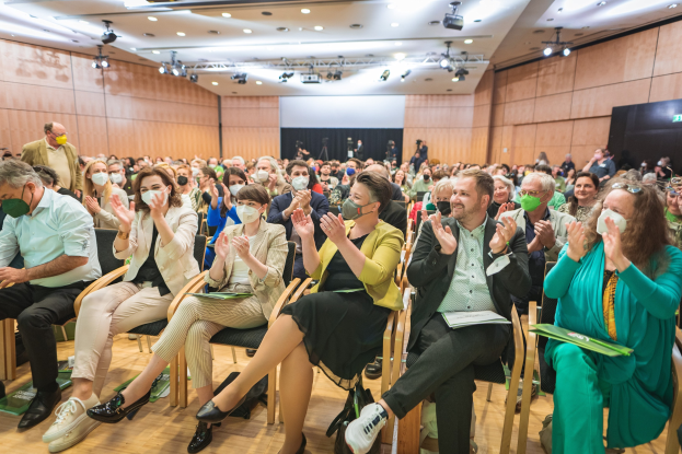 Eine Gruppe von Menschen in Stühlen klatschend, einige tragen Masken, mit Taschen auf dem Boden, vor einem Publikum bei einer Coronavirus-Konferenz mit einem Bildschirm und Deckenlampen im Hintergrund.