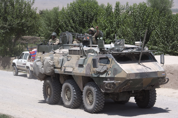Gepanzerter Personentransporter und Lastwagen fahren auf einer Schotterstraße mit bewaffneten Personen in Helmen, umgeben von Bäumen, Hügeln und einem klaren Himmel.