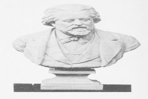 Ein Schwarz-Weiß-Bildnis eines bärtigen Mannes, der mutmaßlich Karl Marx ist, in Anzug und Krawatte, mit ernster Miene und vor der Brust verschränkten Händen.