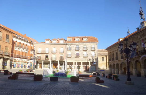Ein Stadtplatz mit einem zentralen Brunnen, umgeben von Bänken, Topfpflanzen, Straßenlaternen und -pfosten sowie einem Uhrenturm und Gebäuden im Hintergrund bei klarem blauem Himmel.