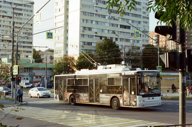 Eine Stadtstraße mit einem Bus, der an hohen Gebäuden, Bäumen, Laternen, Verkehrssignalen und Schildern vorbeifährt, mit Fußgängern auf dem Gehweg, einem Geländer im Vordergrund und einem klaren blauen Himmel.