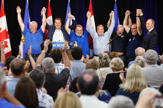 Eine Gruppe von Menschen, darunter der Premierminister von Ontario Doug Ford und seine Unterstützer, steht an einem Tisch mit einer Tafel, auf der "Ontario Premier Doug Ford and his supporters at a campaign event in Ottawa, Ontario, Canada, on Tuesday, October 29, 2019" steht, einem Mikrofon und Flaggen, vor einer Menge, von der einige jubelnd die Hände heben, vor einem Vorhang in einer offiziellen Umgebung.