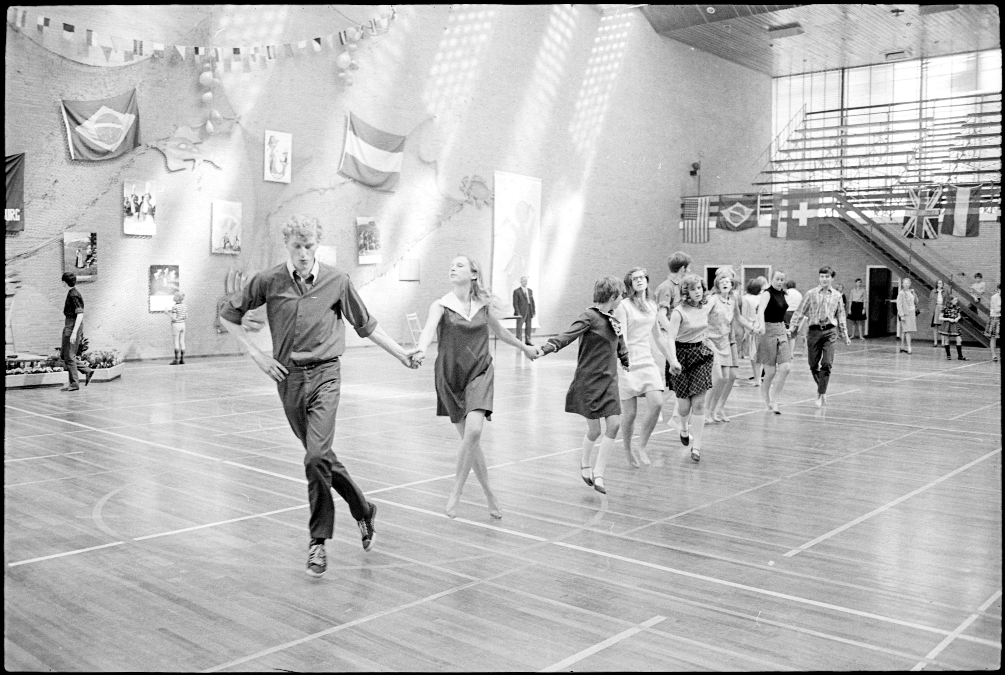 Schwarz-weiß-Foto von Menschen, die in einem Turnsaal tanzen, mit Foto-Rahmen und Flaggen an der Wand, Treppe auf der rechten Seite.