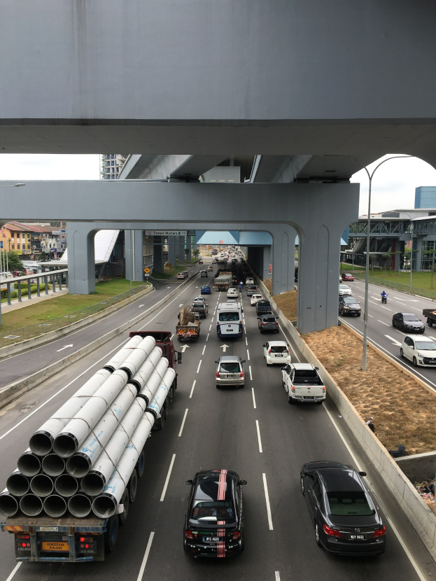 Eine stark befahrene Autobahn mit Schwerlastverkehr unter einer Brücke, umgeben von Gras, Bäumen, Gebäuden, Strommasten und Straßenlaternen unter einem Himmel.