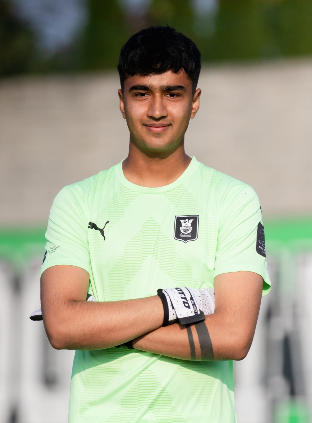 Ein junger Mann in einem grünen Hemd steht auf einem Fußballfeld mit verschränkten Armen und lächelt, wahrscheinlich ein Torwart, mit einem unscharfen Hintergrund, der den Fokus auf ihn legt.