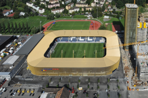 Luftaufnahme eines Fußballstadions im Zentrum einer Stadt, umgeben von Gebäuden, Bäumen, Straßen, Fahrzeugen, Masten und Kränen, mit saftigem Grün.