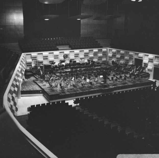 Ein Schwarz-Weiß-Foto eines großen Auditoriums mit einer Bühne in der Mitte, umgeben von Stühlen und beleuchtet von Deckenlampen, auf der ein paar Musiker spielen.