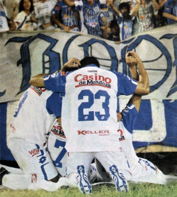 Eine Gruppe von Fußballspielern feiert ein Tor auf dem Feld vor einer jubelnden Menge, im Hintergrund ist ein Banner mit Text zu sehen.