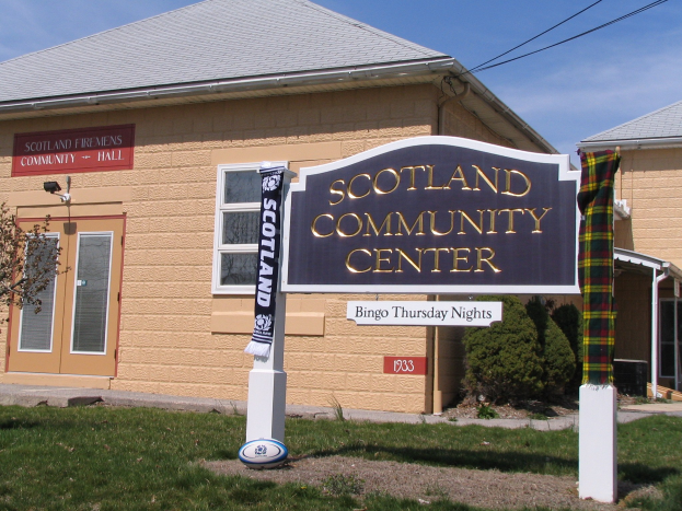 Das Scotland Community Center-Gebäude mit einem Namensschild, umgeben von Gras, einem Baum, Pflanzen, einem Ball, sichtbaren Drähten und einem bewölkten Himmel.