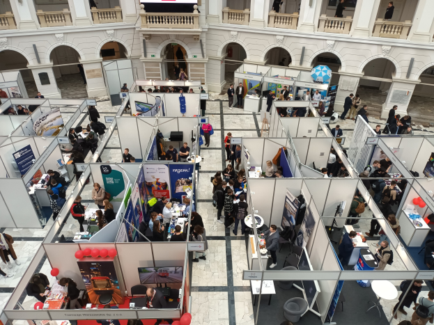 Eine große Gruppe von Menschen auf einer internationalen Handelsmesse mit Ständen, Bannern, Tischen, Stühlen, Luftballons und einem Gebäude mit Bögen, Säulen, Geländern und einem Bildschirm im Hintergrund.