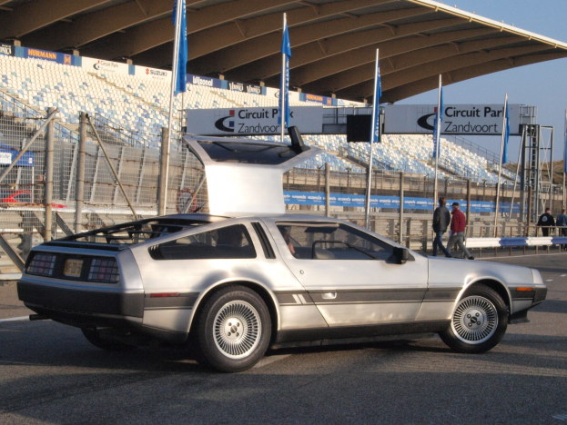 Ein silberner DeLorean DMC-12 fährt auf einer Straße vorbei an Menschen, Fahnen, Schildern, Geländern, Stühlen und einem Schuppen, mit Bäumen und einem klaren blauen Himmel im Hintergrund.