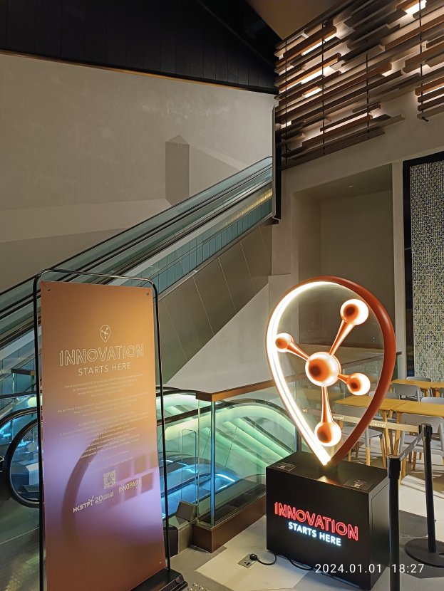 Großes Schild in einem Gebäude mit einer Rolltreppe im Hintergrund, Tische und Stühle auf der rechten Seite und eine Tafel mit der Aufschrift "Innovation Starts Here" auf der linken Seite, die auf die HSTP20 Expo hinweist.