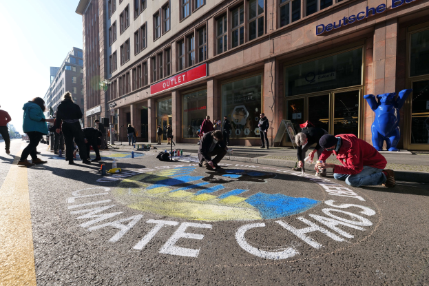 Menschen sitzen vor einem Gebäude mit Fenstern und Namensschildern auf dem Boden, umgeben von Flaschen und anderen Gegenständen, während sie an einer Klimawandel-Demonstration in Berlin teilnehmen, mit Bäumen und einem klaren blauen Himmel im Hintergrund.