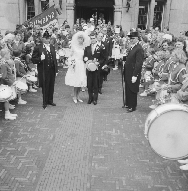 Schwarzes und weißes Foto einer Braut in einem weißen Kleid und Schleier und eines Bräutigams mit einem Blumenstrauß, der eine Straße entlang geht, vorbei an einer Menge mit einigen, die Musikinstrumente halten, mit einem Banner und einem Gebäude im Hintergrund.