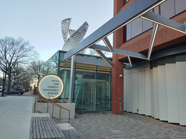 Eintritt zum Faith and Liberty Discovery Center mit Glaswänden, einem Namenbrett, einer Bank, Metallrahmen, Bäumen, Fahrzeugen, Pfählen und einem bewölkten Himmel.