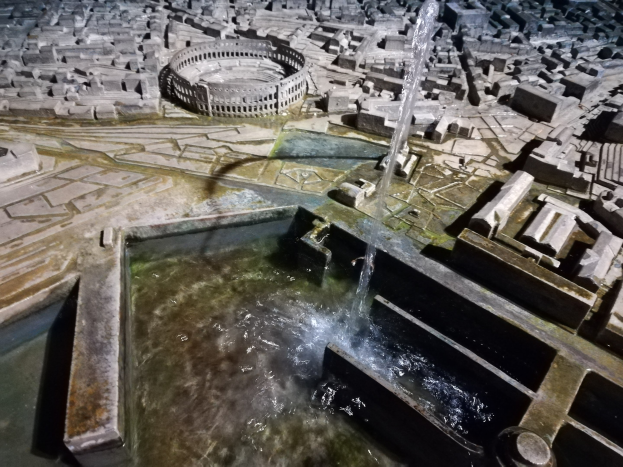 Ein Miniaturstadtmodell mit einem zentralen Springbrunnen mit Wasserfall, umgeben von Gebäuden und einer Wasserfläche.