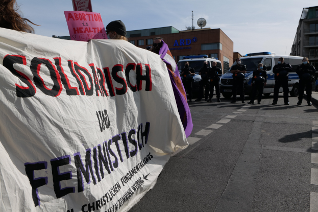 Eine Gruppe von Menschen marschiert auf einer Straße, hält ein Banner mit der Aufschrift "Solidarität und Feminismus" und fährt mit geparkten Fahrzeugen und Gebäuden im Hintergrund unter einem klaren blauen Himmel.