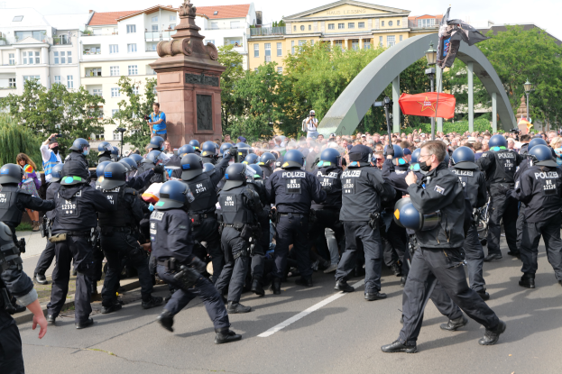 Eine Gruppe von Polizisten in Einsatzausrüstung, mit Helmen, geht eine Straße entlang, umgeben von einer Menge, mit Bäumen, Gebäuden, Laternenmasten, einem Bogen, einer Statue auf einem Sockel und einer Fahne mit einem Mast im Hintergrund unter einem bewölkten Himmel.