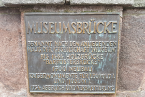 Eine dunkle Plakette an einem steinernen Gebäude mit der fettgedruckten Schrift 'Museumsbrucke'.