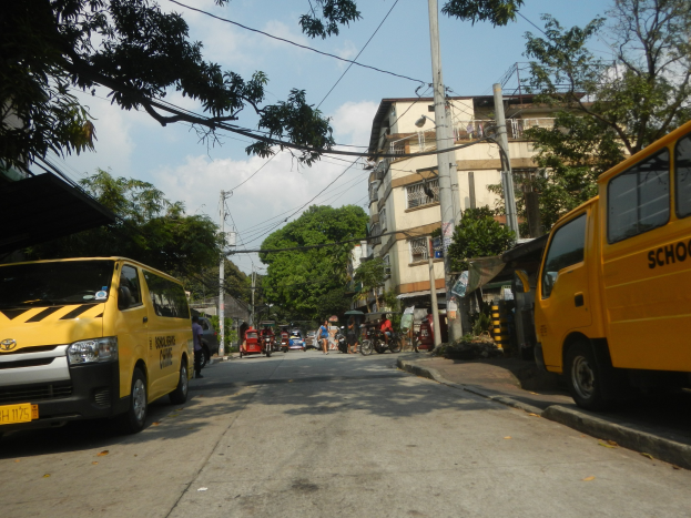Ein gelber Schulbus fährt eine Straße entlang, die von hohen Gebäuden, Bäumen und Strommasten mit Drähten gesäumt ist. Menschen fahren mit Fahrrädern und anderen Fahrzeugen auf der Straße, unter einem bewölkten Himmel.