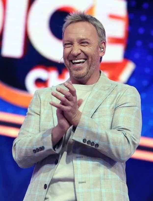 Ein lachender Mann in einem Anzug, der auf einer Bühne klatscht, mit einem 'The Voice'-Schild im Hintergrund.