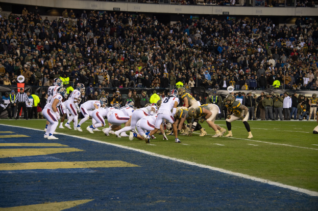 Ein Füssballspiel im Gange mit Spielern in Sportbekleidung und Helmen auf dem Feld, Zuschauern, Geländern und Tafeln mit Text im Hintergrund.