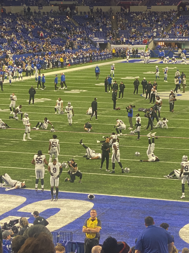 Ein Fußballspiel wird in einem Stadion mit Zuschauern auf dem Rasen und einigen auf Stühlen sitzenden Personen gespielt, während eine Person eine Kamera hält, was auf eine Live-Übertragung hinweist.
