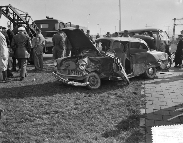 Eine Schwarz-Weiß-Szene eines verunglückten Autos am Straßenrand mit vielen Menschen drumherum, einige tragen Mützen, und eine Person hält ein Fahrrad, mit Laternenmasten und einem klaren Himmel im Hintergrund.