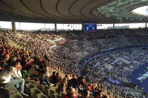 Große Menschenmenge in einem Stadion bei einem Fußballspiel, mit einer Bühne rechts, Fahnen, Stangen, einem Bildschirm und dem Allianz Stadion in München, Deutschland im Hintergrund.