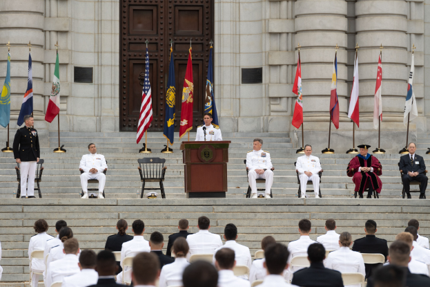 Eine Gruppe von Menschen in weißen Uniformen sitzt auf Stufen vor einem Podium, das von Fahnen flankiert ist, mit einem großen Gebäude und einer Tür im Hintergrund.