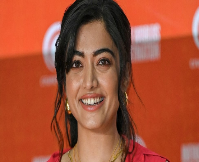 Eine Frau in einem roten Kleid mit einem eleganten Dutt und dezentem Make-up lächelt in die Kamera, trägt schwarze High Heels und hat im Hintergrund ein hellgelbes Banner mit der Aufschrift "Sonam Kapoors schönste Looks vom Cannes Filmfestival".