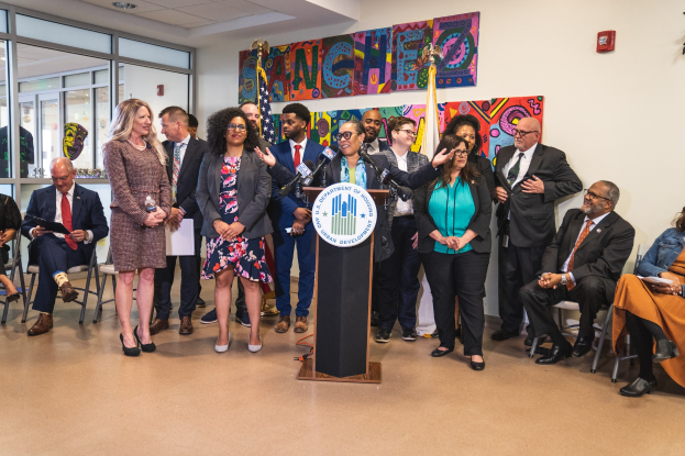 Gruppe von Menschen vor einem Podium mit einem Mikrofon und einer Tafel mit der Aufschrift "Sancho Stadtrat tritt dem Stadtrat bei", mit sitzenden und stehenden Teilnehmern, Flaggen, einem Feuerlöscher und Deckenbeleuchtung.