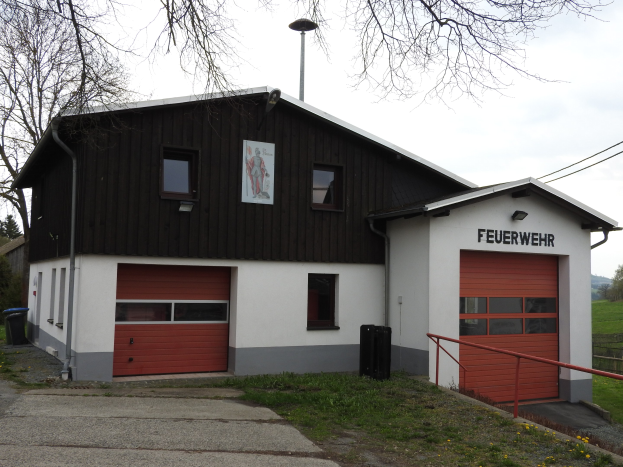 Feuerwache mit einem roten und weißen Gebäude, Straße, Gras, gelben Blumen und Bäumen im Hintergrund.