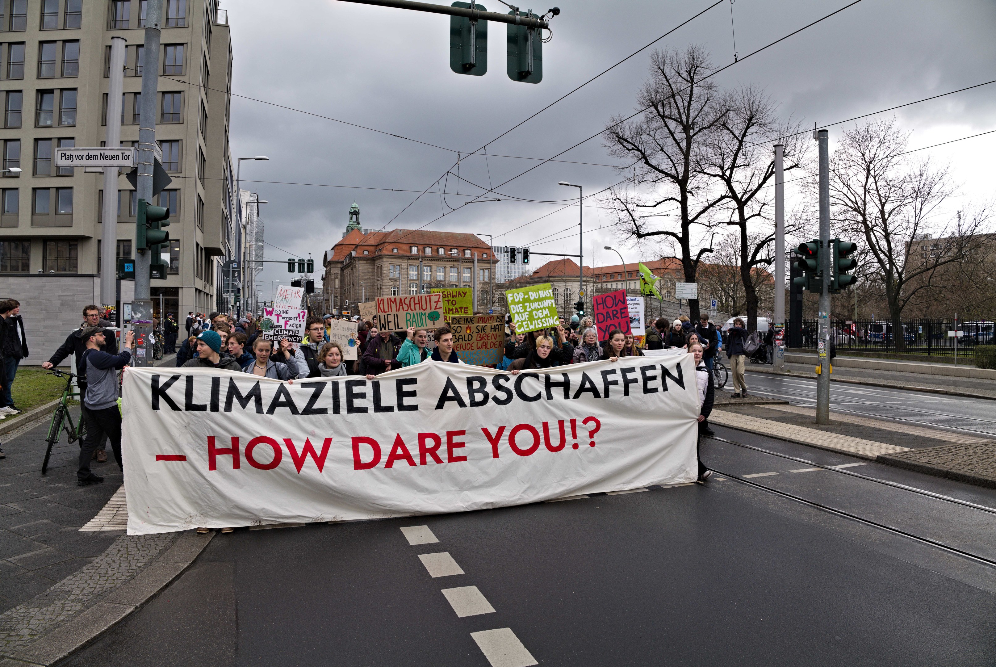 Gruppe von Menschen marschiert mit einem Banner "Klimaziele Abschaffen - Wie wagst du es?" die Straße entlang, mit einem Radfahrer im Vordergrund und Gebäuden, Bäumen, Strommasten, Verkehrszeichen und einem klaren blauen Himmel im Hintergrund.