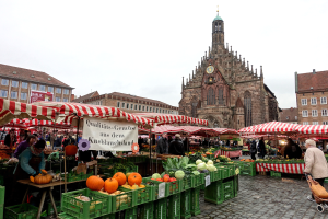 Ein lebendiger Markt in Nürnberg, Deutschland, voller verschiedener Früchte und Gemüse, Menschen mit Taschen und Zelten und im Hintergrund Gebäude und ein Uhrenturm unter einem klaren Himmel.