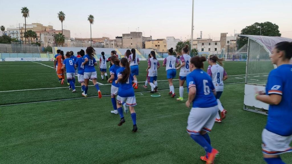 Eine Gruppe von Frauen beim Fußballspielen auf einem Outdoor-Feld mit Gebäuden, Bäumen und einem Torpfosten im Hintergrund.