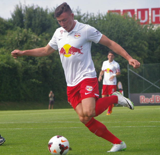 Ein Profifußballer tritt einen Ball auf einem grünen Feld mit "RB Leipzig" im Hintergrund.