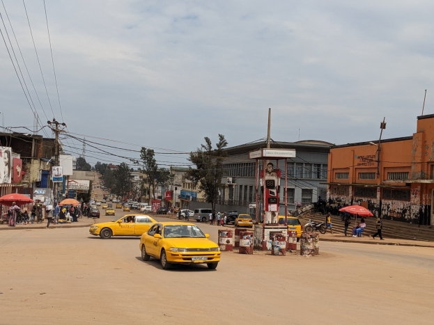 Ein gelbes Taxi fährt auf einer belebten Stadtstraße mit Gebäuden an den Seiten, während Fußgänger auf dem Gehweg gehen, einige halten Schirme, unter einem bewölkten Himmel mit sichtbaren Strommasten und -drähten.