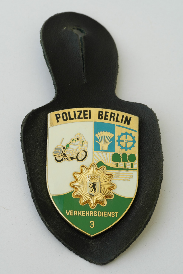 Ein schildförmiges Polizeibadgebnis mit "Polizei Berlin" in schwarzer Schrift auf einem blauen Hintergrund mit einem weißen Stern und Rahmen, das auf einer Oberfläche ruht.