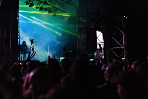 Menge bei einem Konzert mit Musikern auf der Bühne, die Instrumente spielen, unter Bühnenbeleuchtung, mit einem Banner und Metallrahmen im Hintergrund.