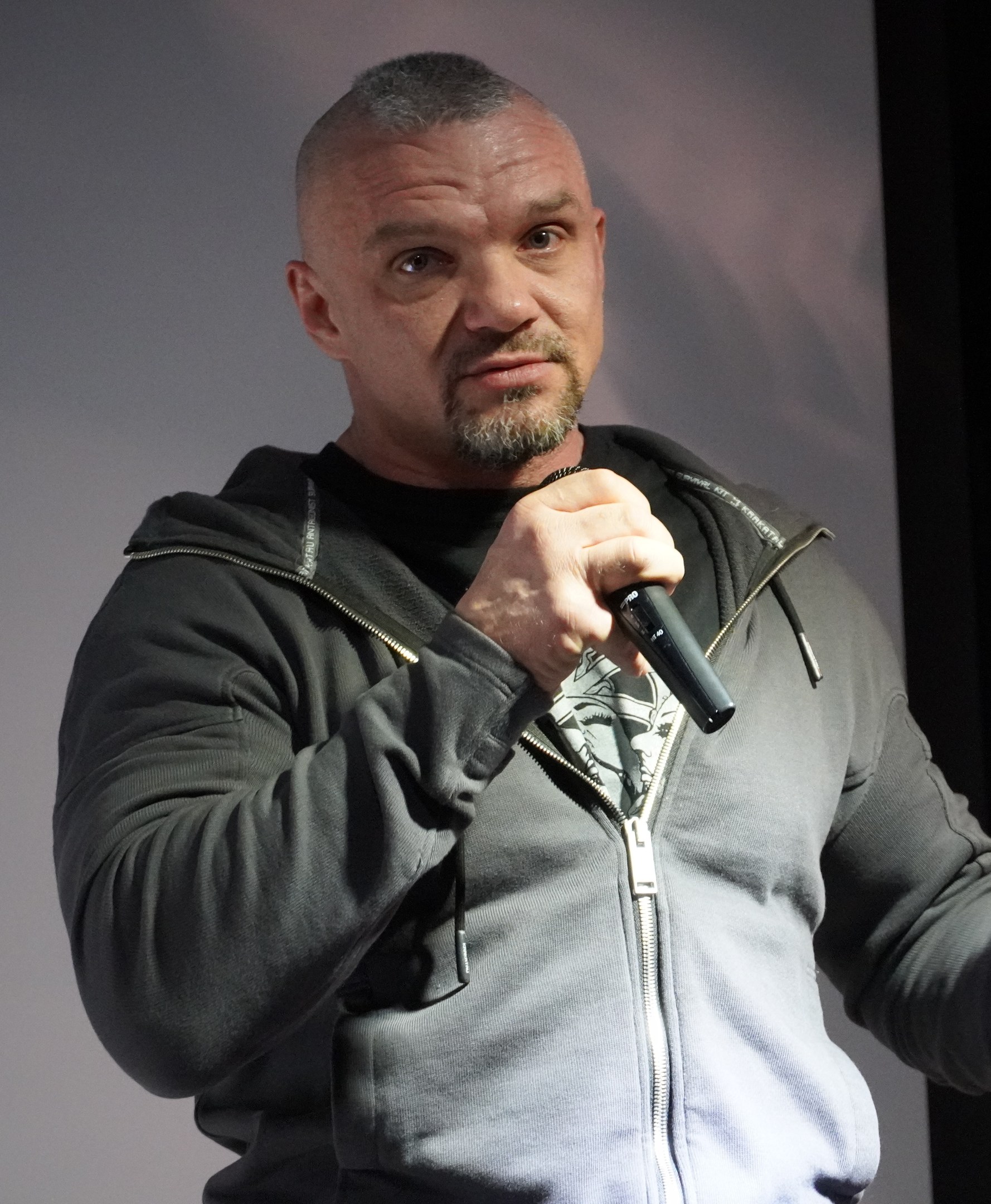 Ein Mann in einer schwarzen Kapuzenjacke und einem schwarzen Anzug mit einem wei├čem Hemd, der ein Mikrofon haltend, selbstbewusst vor einer Wand steht und wie ein Komiker mit zurückgek├Ąmmtem Haar wirkt.