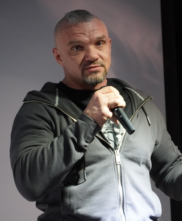 Ein Mann in einer schwarzen Kapuzenjacke und einem schwarzen Anzug mit einem wei├čem Hemd, der ein Mikrofon haltend, selbstbewusst vor einer Wand steht und wie ein Komiker mit zurückgek├Ąmmtem Haar wirkt.