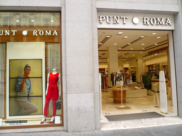 Vorderfront von Punt Roma in Mailand, Italien, mit Schaufensterpuppen in Kleidern, beleuchtet von Deckenlampen, mit Text an der Wand und einem Türmatten auf dem Boden.