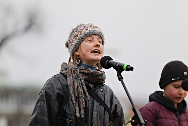 Eine Frau in einer schwarzen Jacke, einem Schal und einer Mütze singt in ein Mikrofon auf der Bühne, neben ihr eine Person in einer maronenfarbenen Jacke und einer schwarzen Mütze, während ein unscharfer Hintergrund die Szenerie bildet.