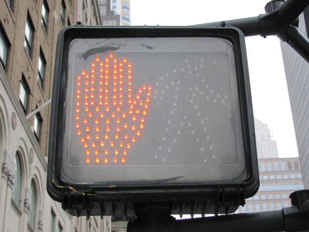 Eine Ampel mit rotem Hand-Symbol in einer städtischen Umgebung mit Gebäuden auf beiden Seiten und einem klaren Himmel darüber.