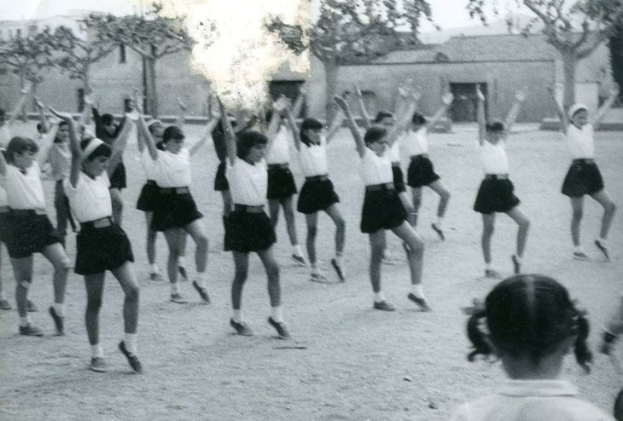 Schwarzes und weißes Foto von jungen Mädchen in Schuluniformen, die auf einer Straße tanzen, die von Gebäuden und Bäumen gesäumt ist, mit einer Person in Schuhen am unteren Rand.